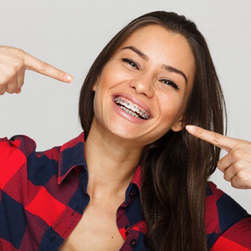 Woman with brown hair in red plaid shirt pointing to her teeth with braces and smiling